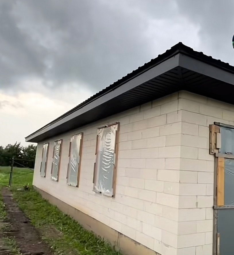 Dom w trakcie budowy z zamontowanym, czarnym dachem i zabezpieczonymi oknami folią, widok z boku budynku przy pochmurnym niebie.