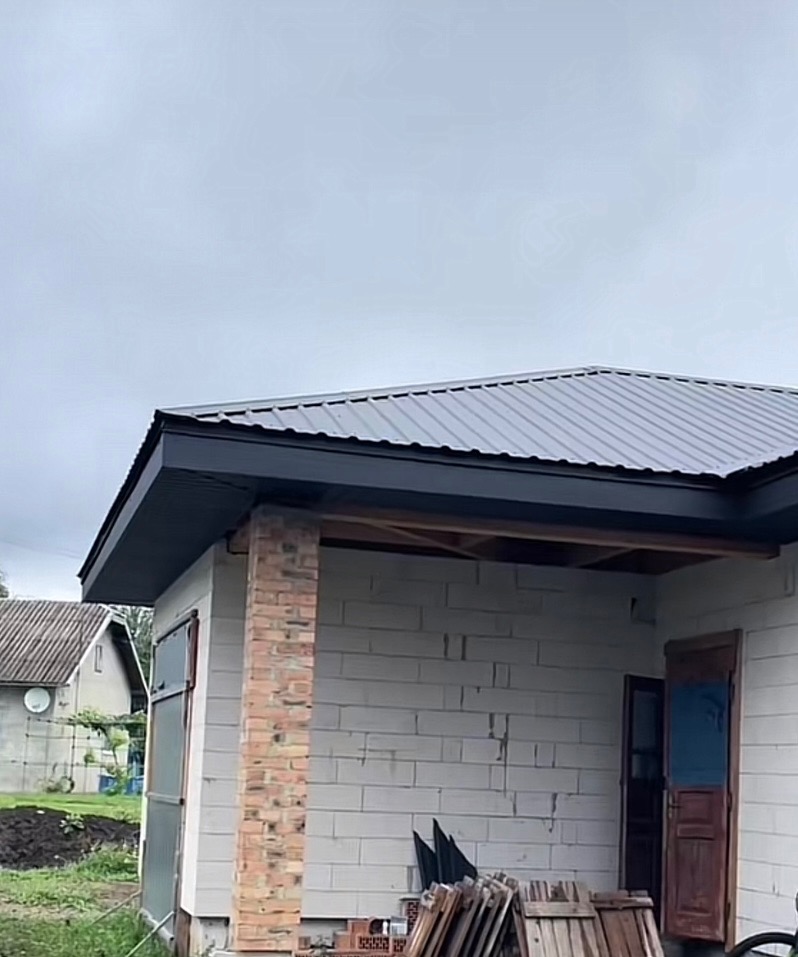 Szary dach z blachy trapezowej z czarnymi rynnami na budynku w trakcie budowy. Widoczny mur z cegły i bloczków. Niebo pochmurne.
