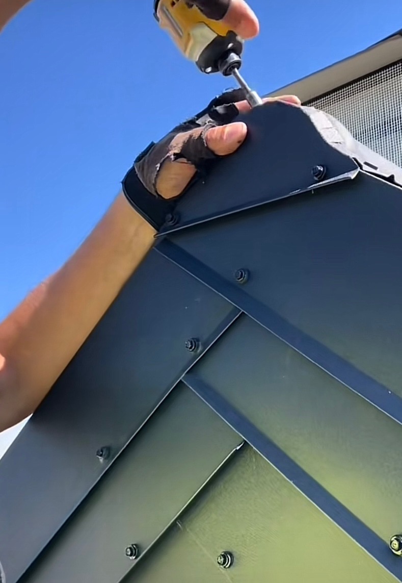 Monterażysta mocuje blachę trapezową do konstrukcji dachu za pomocą wkrętarki. Widoczne połączenia śrubowe i fragment niebieskiego nieba.