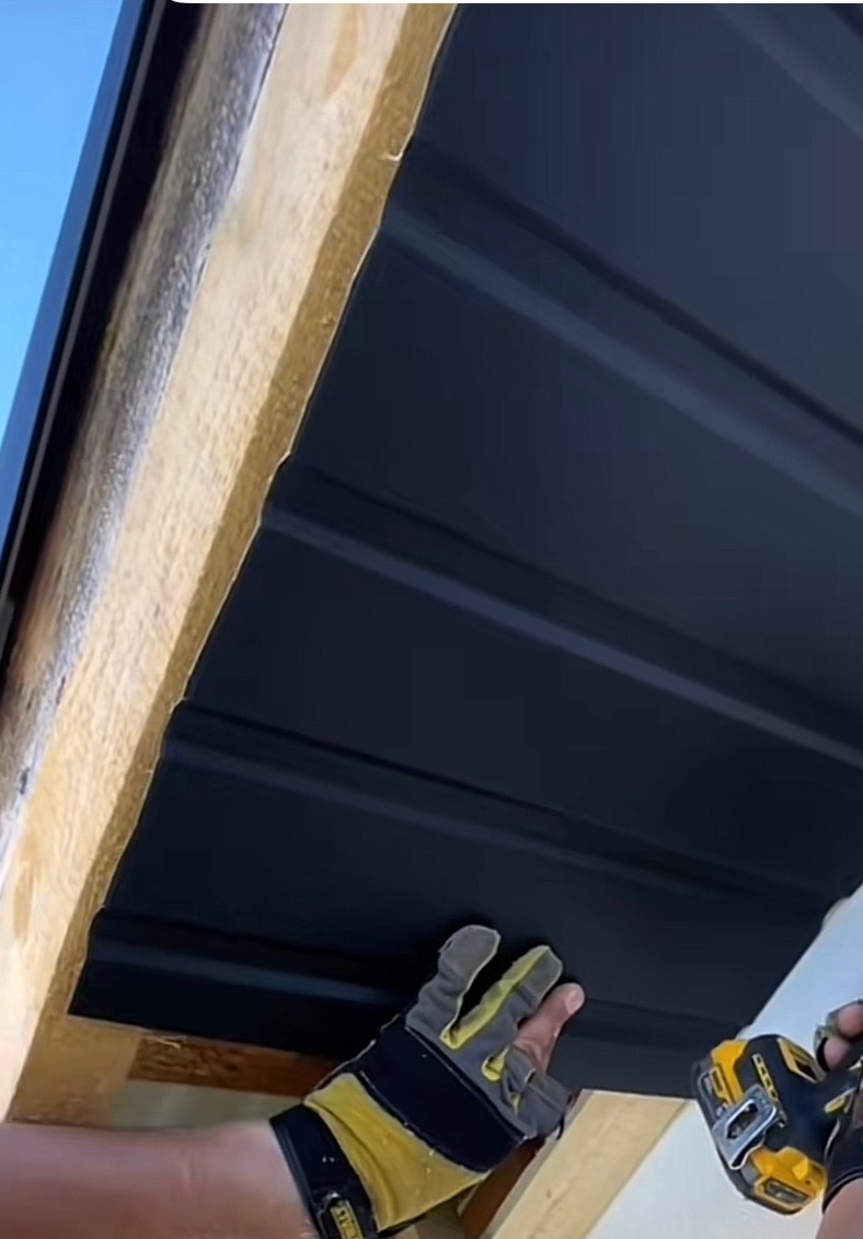 Montaż ciemnej blachy trapezowej na dachu, widoczne drewniane elementy konstrukcyjne i dłoń w rękawicy trzymająca wkrętarkę.