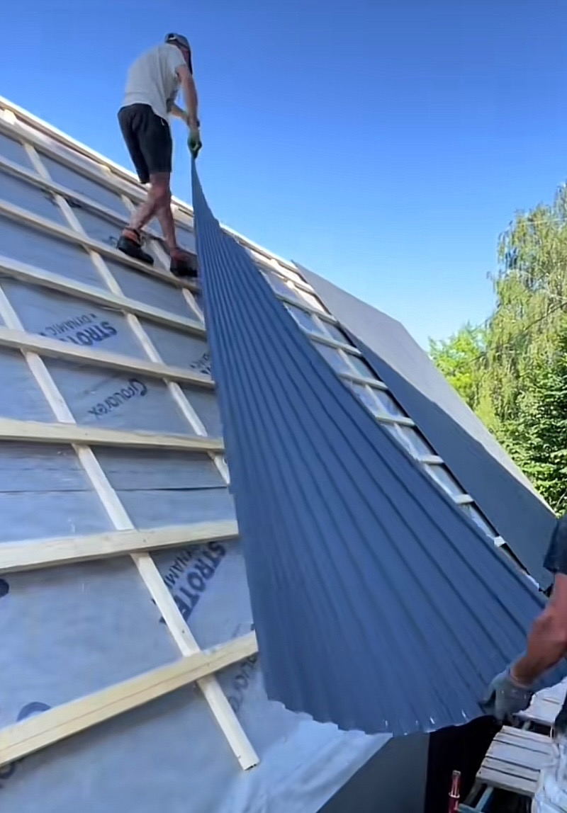 Montaż paneli z blachy na dachu z widoczną drewnianą konstrukcją. Jeden z pracowników trzyma panel, drugi stoi na dachu. Widoczne membrany dachowe.