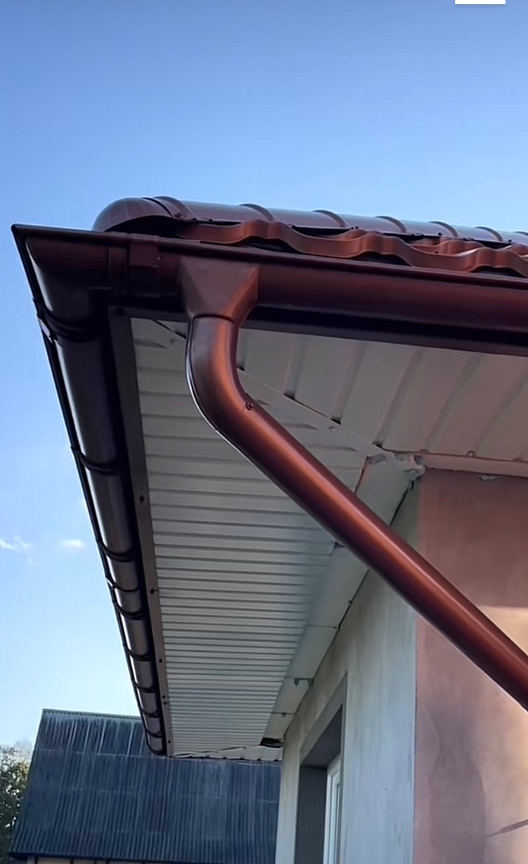 Brązowa rynna z metalowym dachem na tle błękitnego nieba. Widoczny fragment elewacji budynku i okno. Ujęcie z dołu, podkreślające detale.