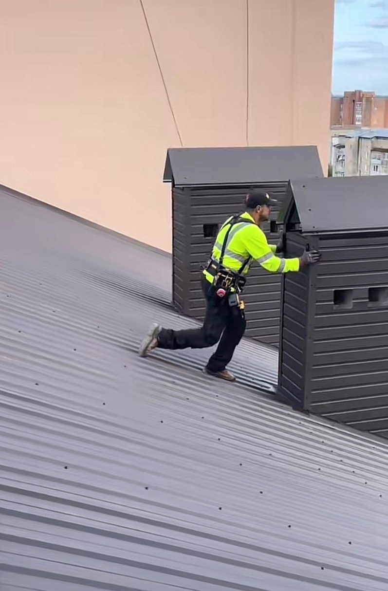 Pracownik w kamizelce na dachu z blachy, montuje szare elementy wentylacyjne. Widok na budynki w tle.