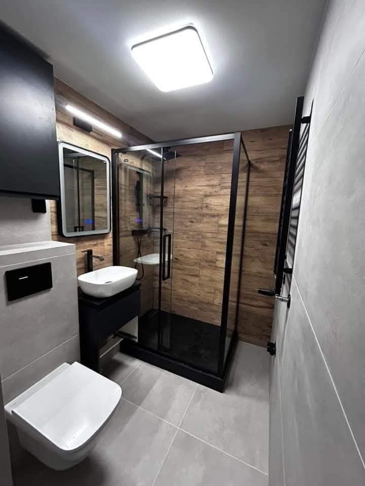 Nowoczesna łazienka z kabiną prysznicową typu walk-in z czarną ramą, umywalką nablatową i toaletą podwieszaną. Ściany wyłożone płytkami imitującymi drewno i szarym gresem.