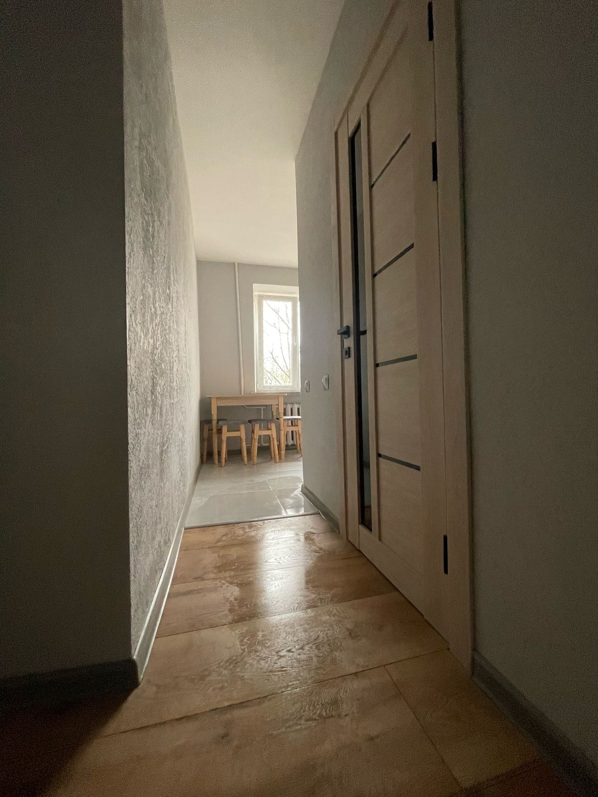 Wykończony korytarz z panelami podłogowymi, szarą ścianą z fakturą i drzwiami z przeszkleniami. W tle widoczny pokój z oknem i drewnianym stołem z krzesłami.