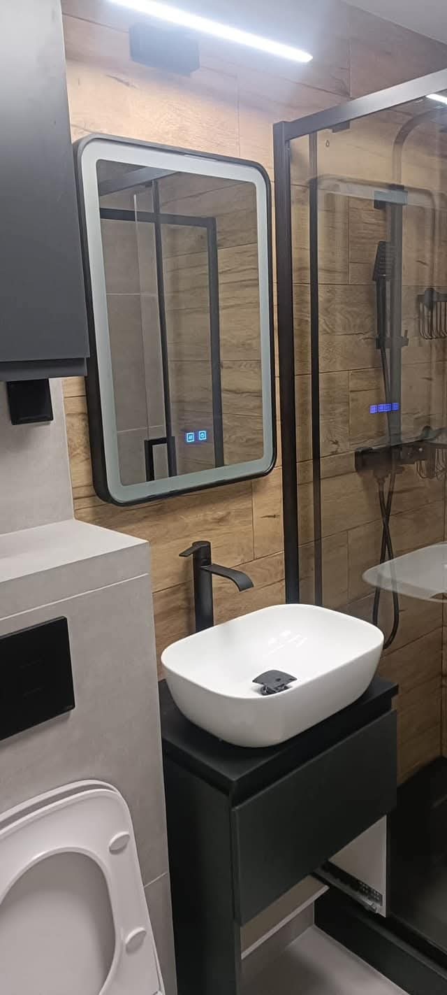 Nowoczesna łazienka z podświetlanym lustrem, białą umywalką nablatową i kabiną prysznicową z czarnymi ramami na tle drewnopodobnych płytek.