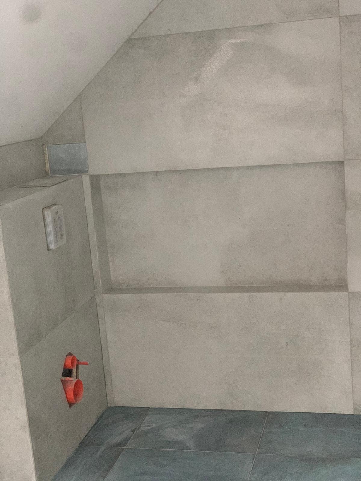 Wykończona ściana łazienki z szarymi, matowymi płytkami. Widoczne fragmenty instalacji wodnej i elektrycznej. Minimalistyczny design.