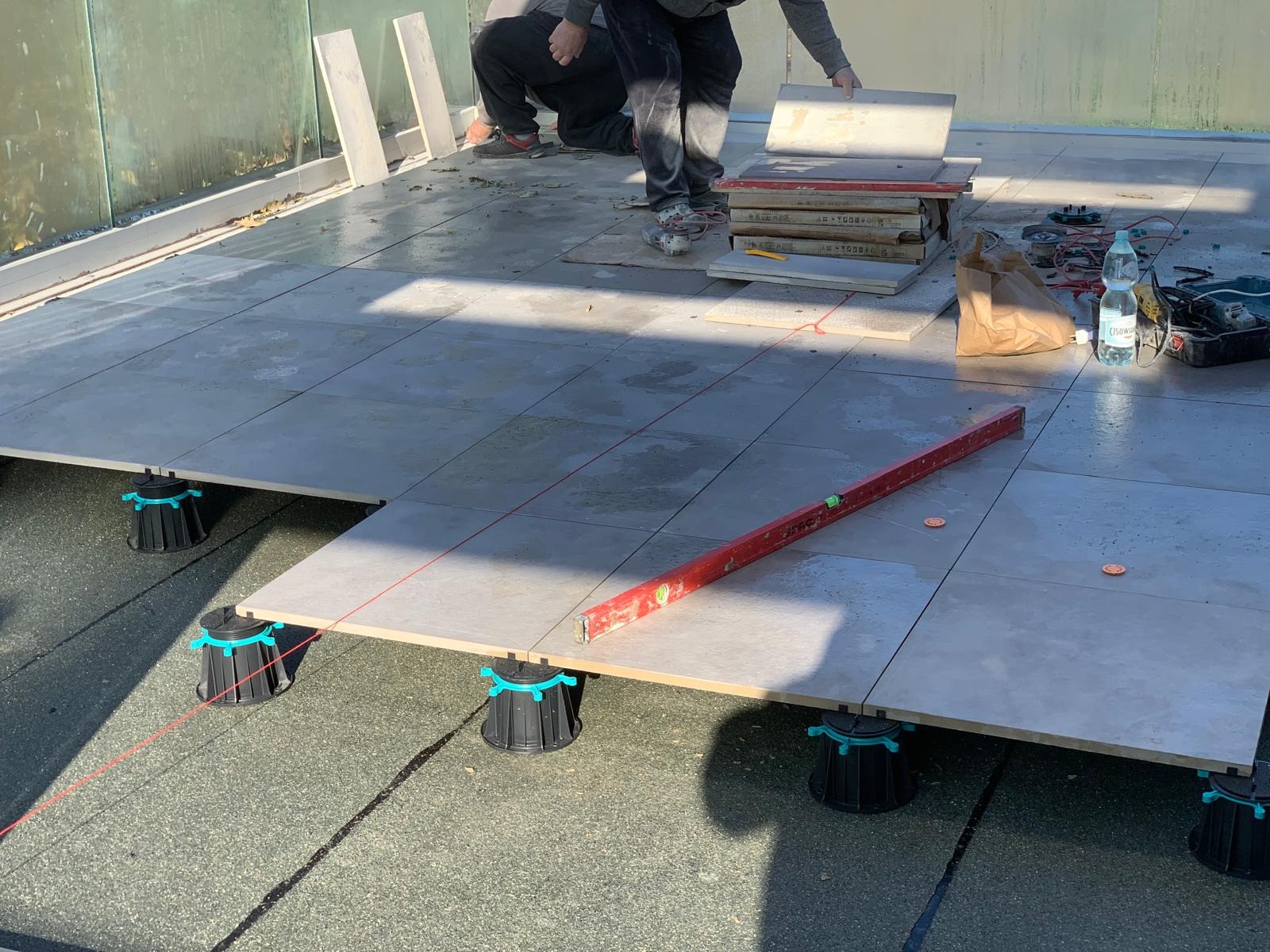 Układanie płytek na tarasie na regulowanych wspornikach. Widoczny poziomica, narzędzia i materiały. Praca w toku, płytki w odcieniach szarości.