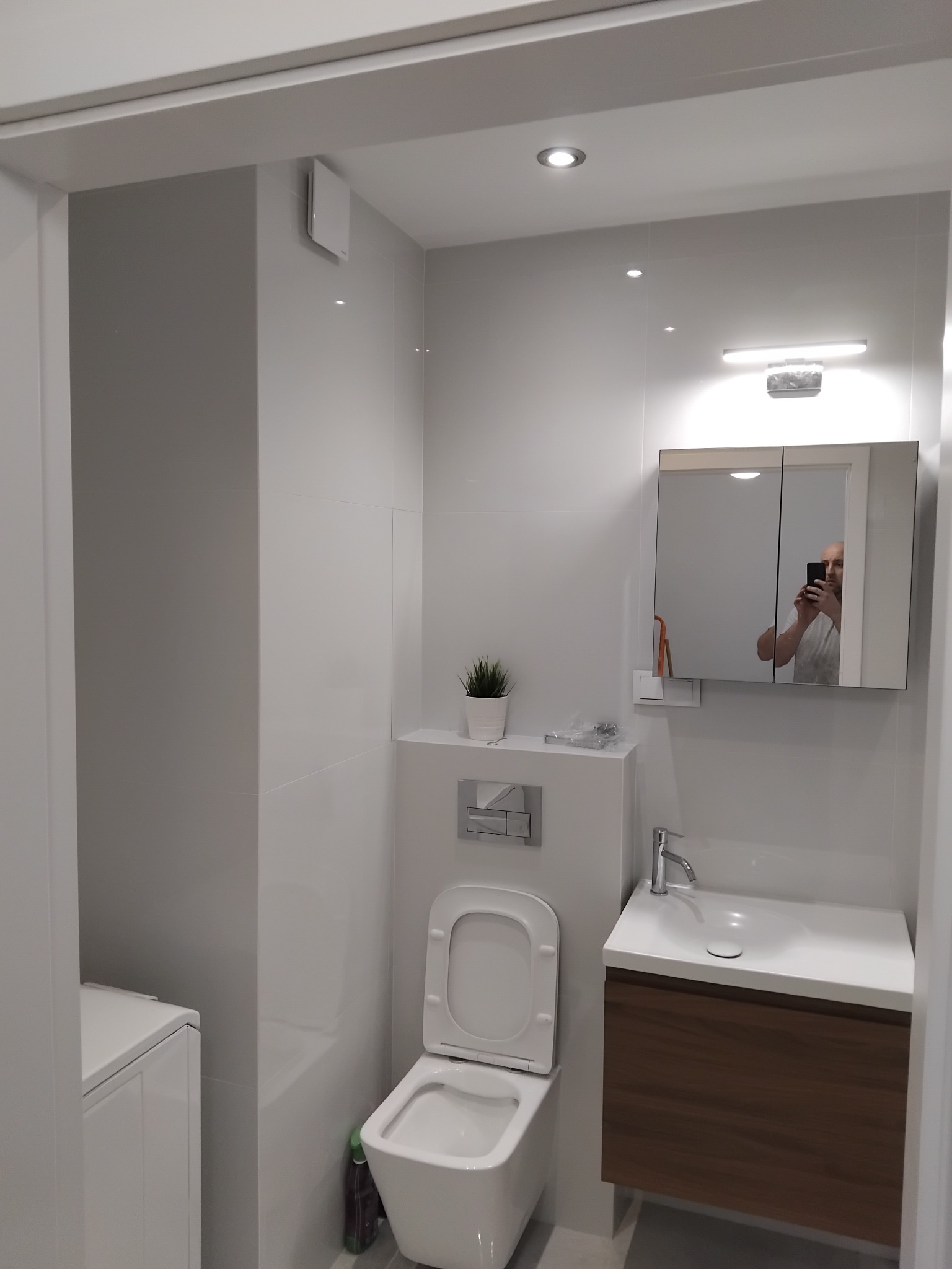 Nowoczesna, jasna łazienka z białymi płytkami, wiszącą toaletą, umywalką z szafką w kolorze drewna i lustrem, w którym odbija się osoba robiąca zdjęcie.