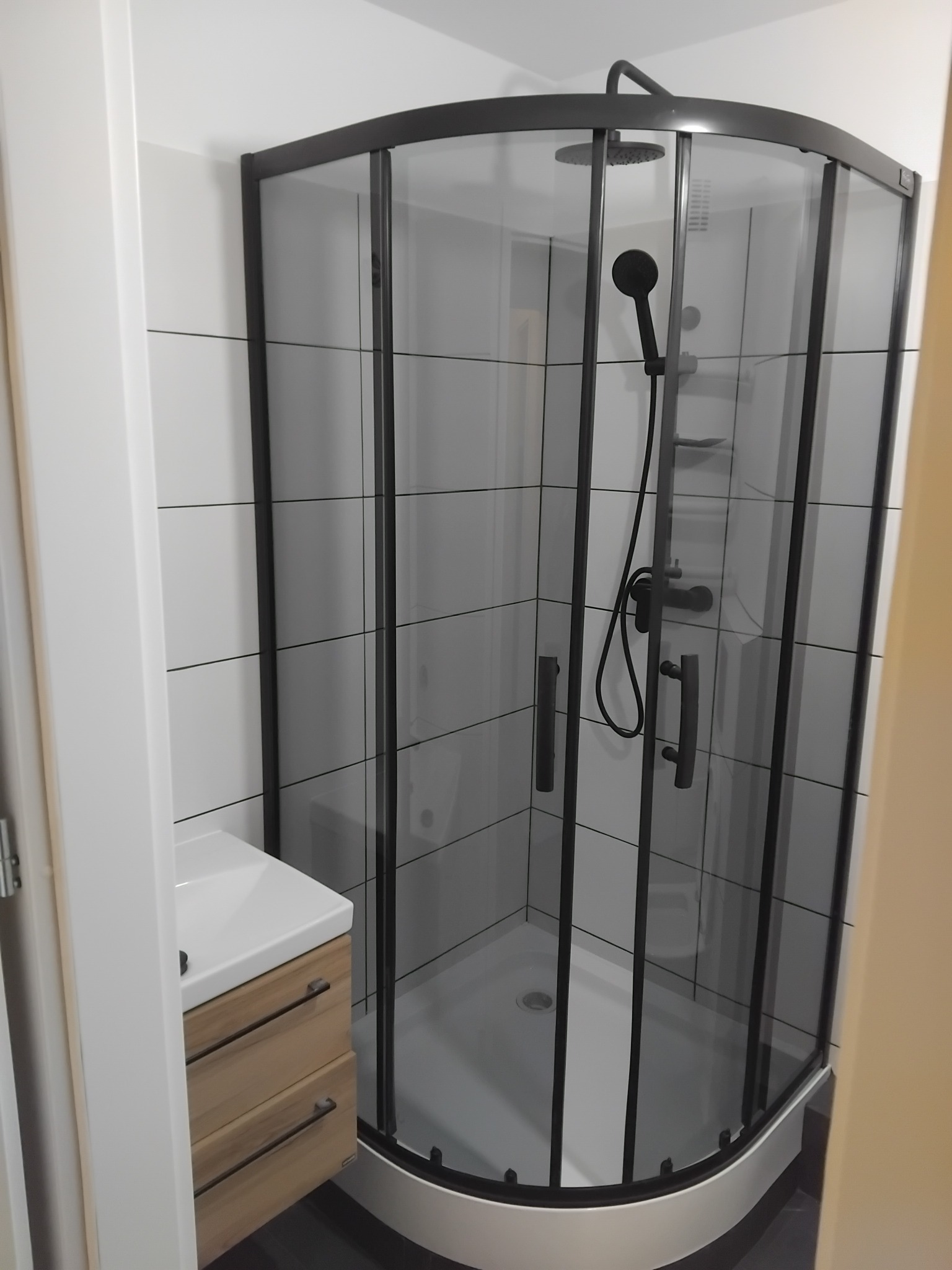 Nowoczesna kabina prysznicowa z czarnymi profilami i armaturą, umywalka z szafką w jasnym drewnie, białe płytki na ścianie. Wykończona łazienka.