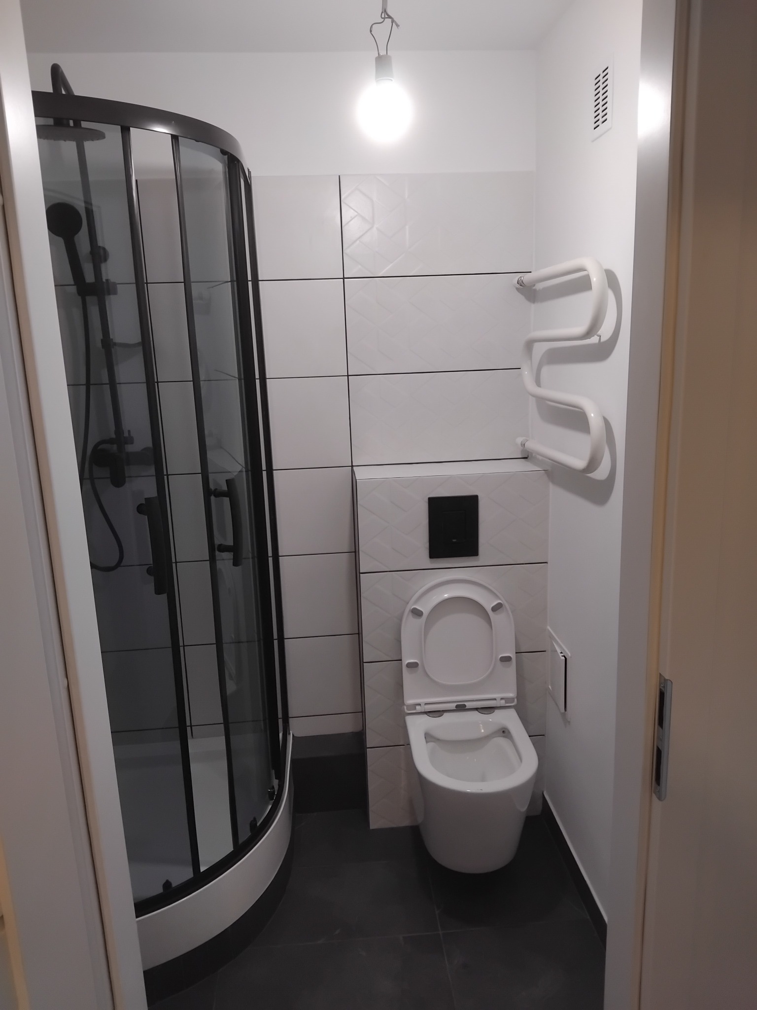 Nowoczesna łazienka z kabiną prysznicową typu walk-in, wiszącą toaletą i grzejnikiem drabinkowym. Białe płytki z subtelnym wzorem i ciemna podłoga tworzą elegancki kontrast.