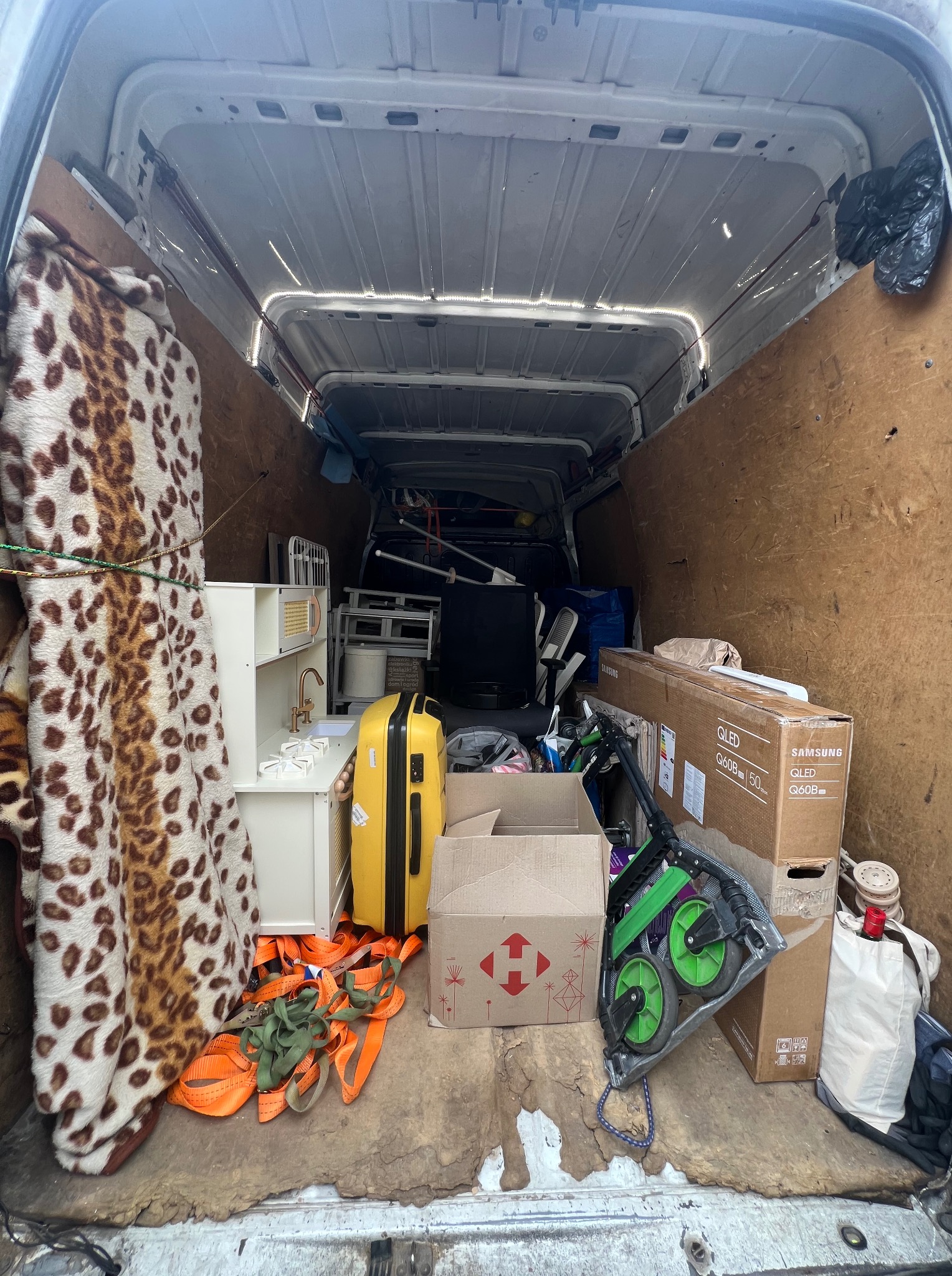 Wnętrze busa wypełnione różnorodnymi przedmiotami gotowymi do transportu: meble dziecięce, walizka, pudła, koc w panterkę oraz zabezpieczenia.