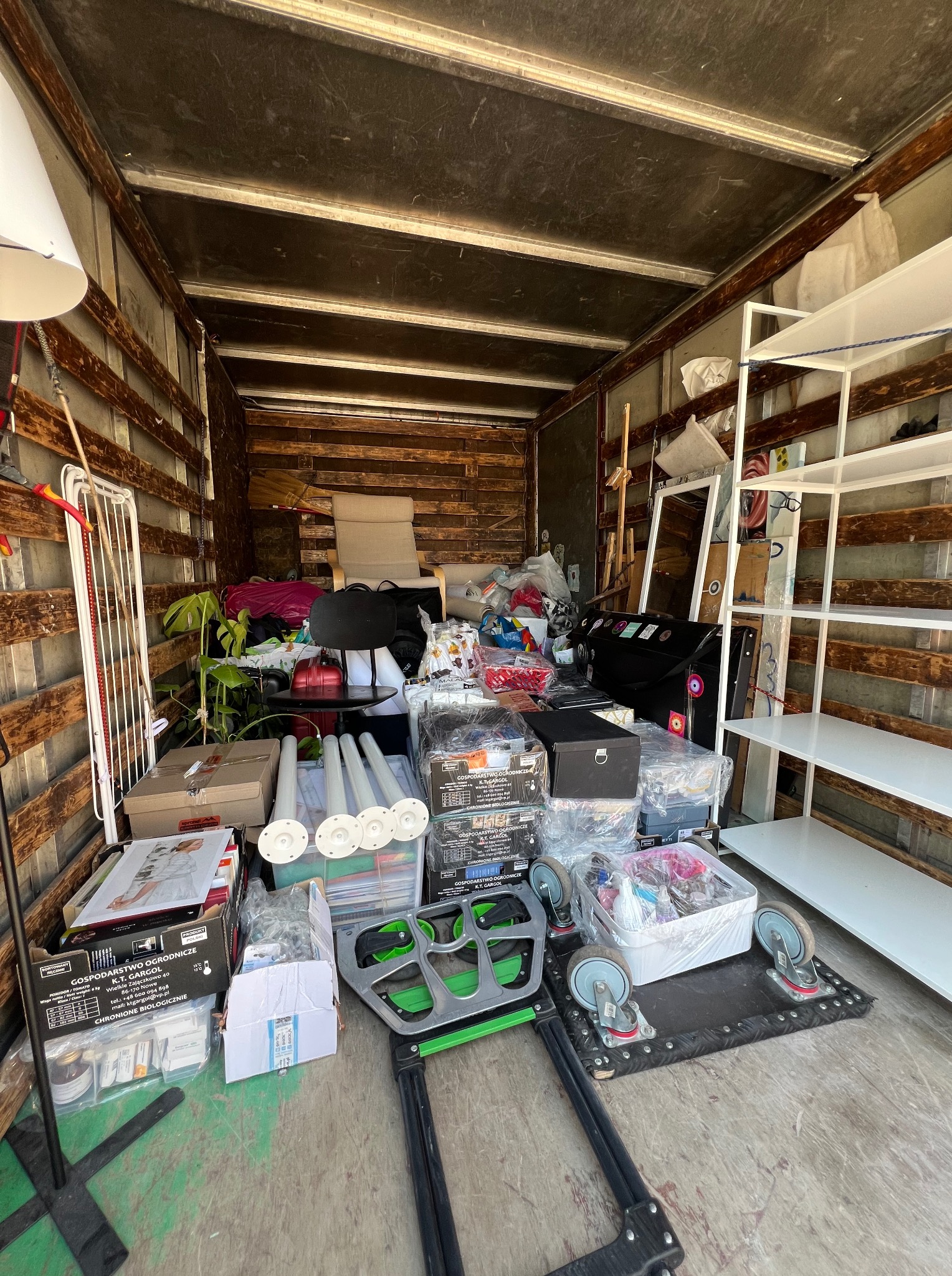 Wnętrze drewnianej furgonetki wypełnionej meblami, roślinami i kartonami, gotowej do transportu. Widoczny wózek transportowy z kółkami.