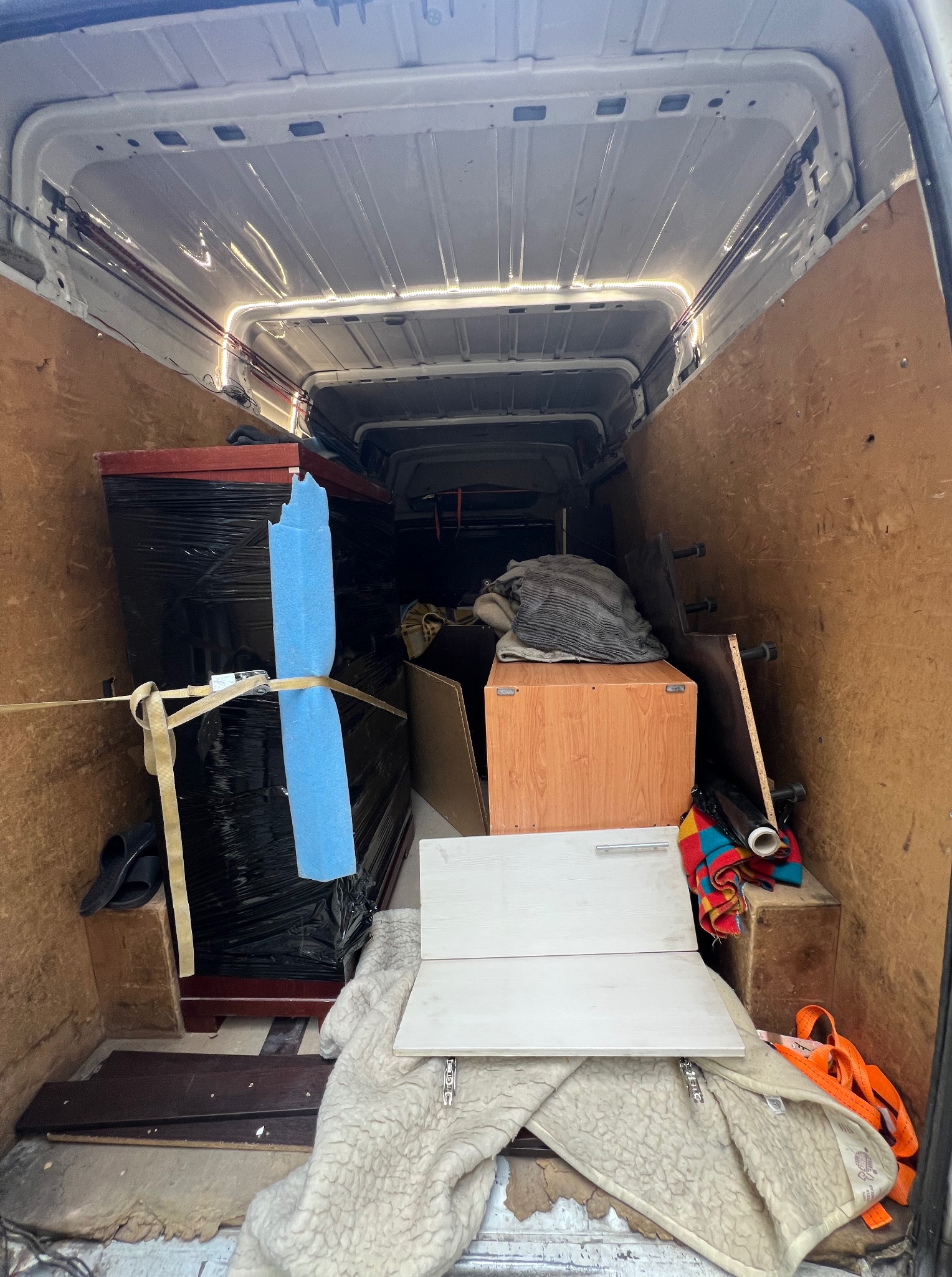 Wnętrze busa z meblami zabezpieczonymi folią i pasami. Widoczne elementy wyposażenia i kartony. Oświetlenie LED wzdłuż sufitu.