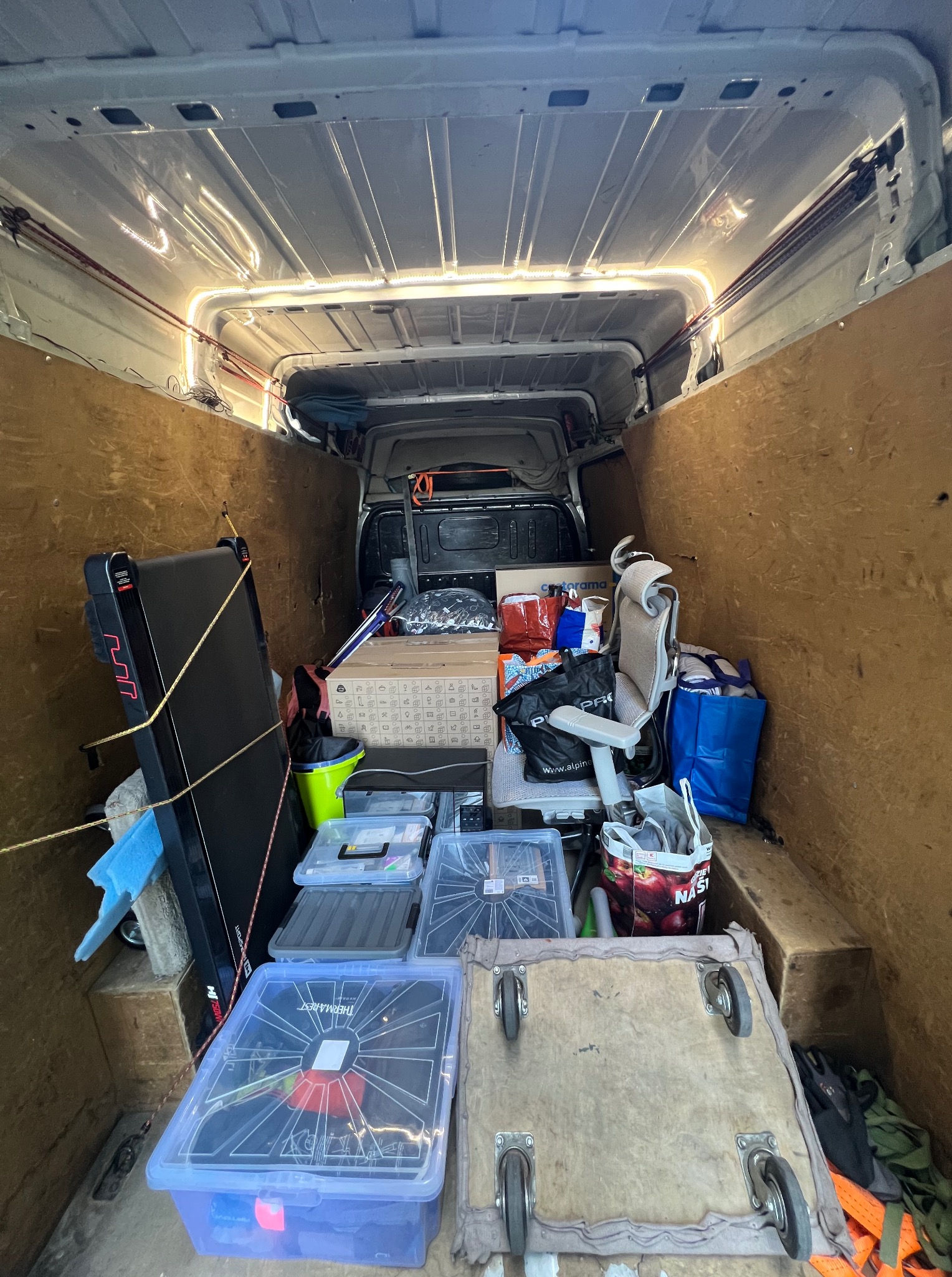 Wnętrze furgonetki wypełnionej różnorodnymi przedmiotami, w tym kartonami, sprzętem sportowym i meblami biurowymi, zabezpieczonymi sznurami. Widok z tyłu pojazdu.