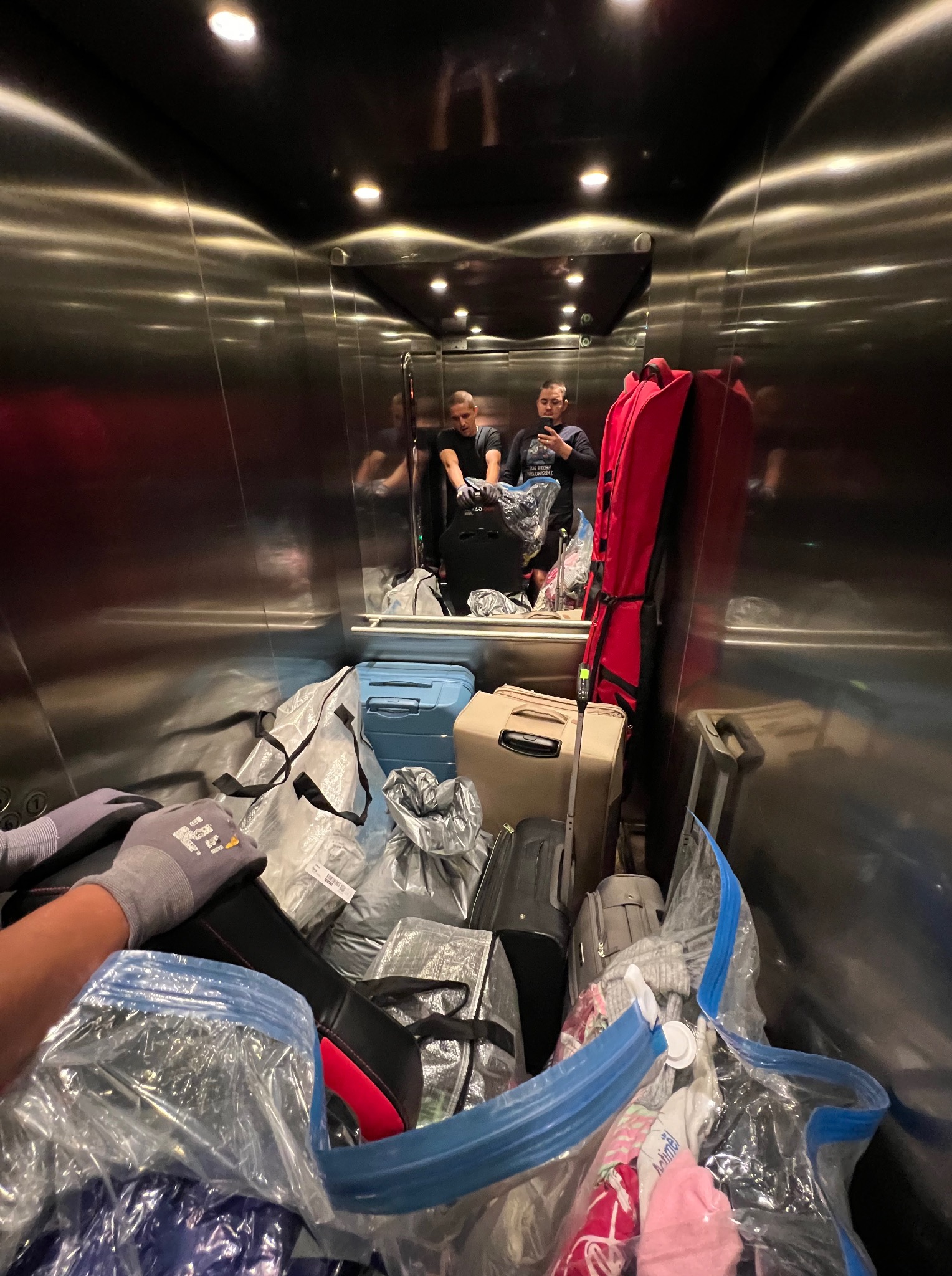 Przepełniona bagażami winda z odbiciem dwóch osób. Walizki, torby w folii i pokrowiec na narty. Widok na załadowaną windę z perspektywy osoby pakującej.
