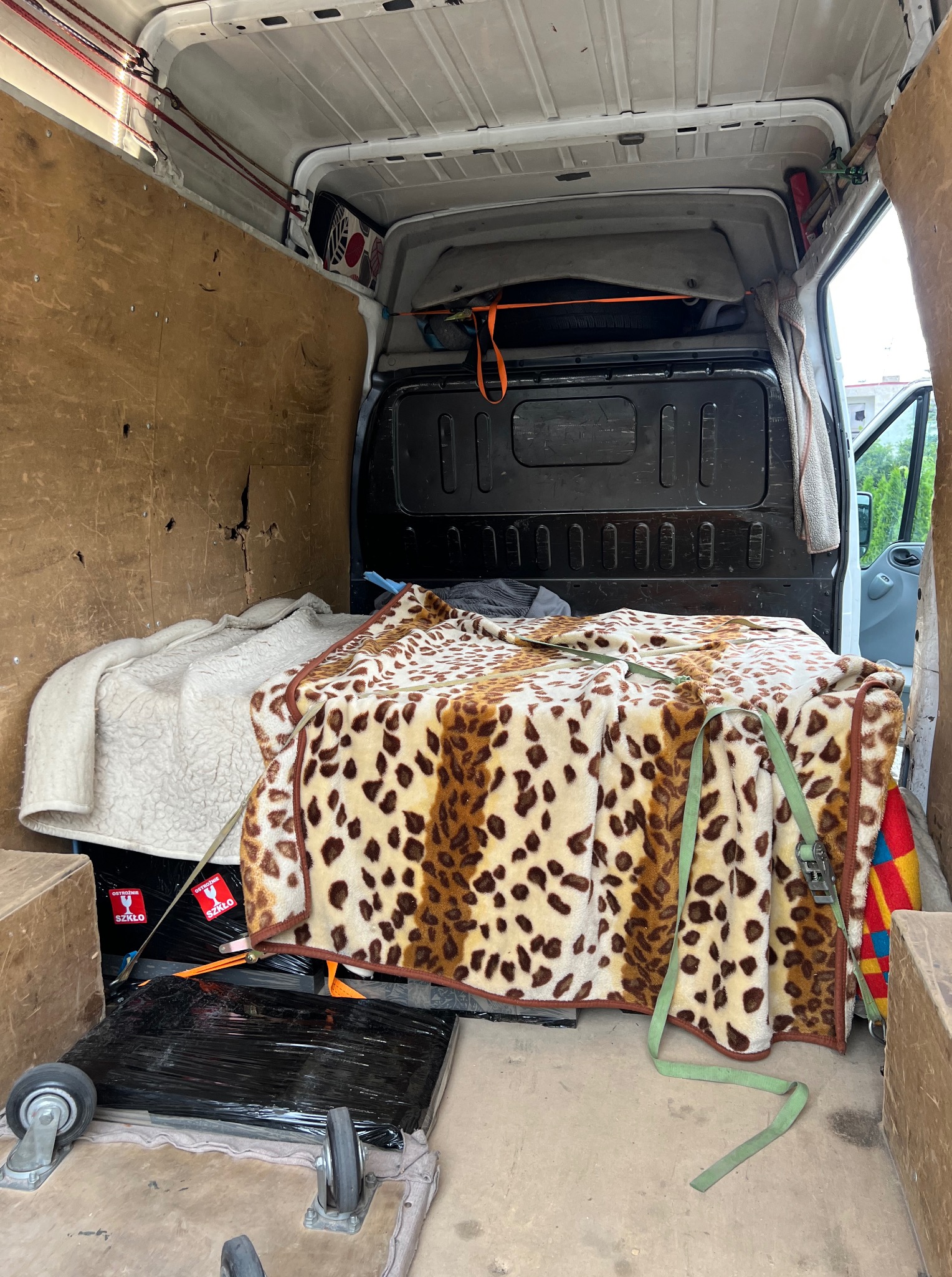 Wnętrze busa z zabezpieczonymi kocami i pasami transportowymi przedmiotami, przygotowanego do przewozu. Widoczne kartony i oznaczenia 'Ostrożnie Szkło'.