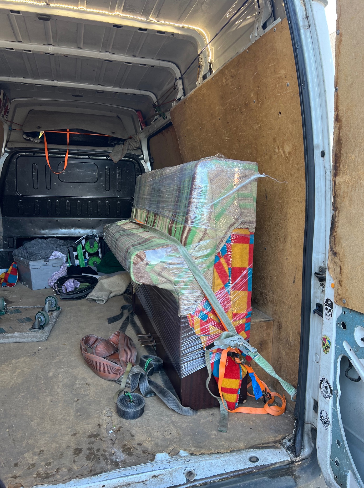 Pianino zabezpieczone folią i pasami transportowymi wewnątrz furgonetki, gotowe do przewozu. Widoczne elementy wyposażenia i naklejki na drzwiach.