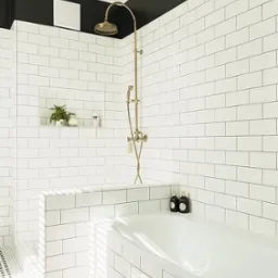 Biała łazienka z wanną obudowaną płytkami, armaturą w kolorze starego złota i dywanem w orientalny wzór. Czarny pas na górze ściany dodaje kontrastu.