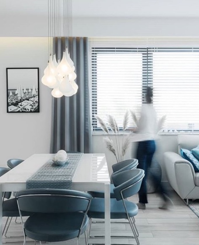 Jasna jadalnia z białym stołem, błękitnymi krzesłami i oryginalną lampą wiszącą. W tle okno z żaluzjami i sylwetka przechodzącej osoby.