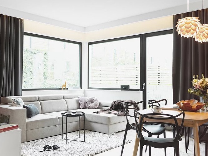 Jasne, nowoczesne wnętrze: narożna sofa, stół z krzesłami, duże okna z widokiem na zieleń. Minimalistyczne oświetlenie i dekoracje tworzą przytulną atmosferę.