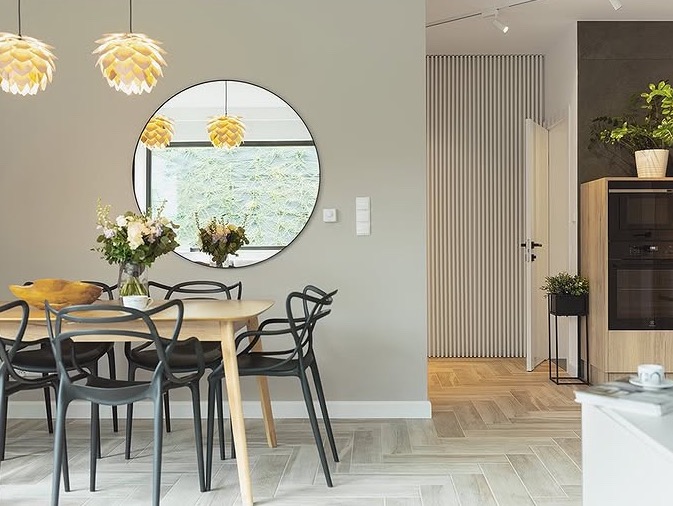 Stylowa jadalnia z okrągłym lustrem, drewnianym stołem i czarnymi krzesłami. Parkiet ułożony w jodełkę, designerskie lampy wiszące. Widok na sąsiednie pomieszczenie.