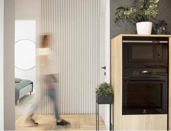Nowoczesne wnętrze: zabudowa kuchenna z piekarnikiem i mikrofalówką, dekoracyjna ścianka z pionowych paneli, osoba w tle. Stylowe wykończenie.