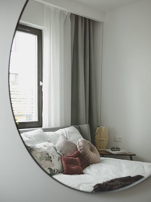 Sypialnia odbita w okrągłym lustrze: łóżko z poduszkami, zasłony w oknie, lampka nocna i okulary na stoliku. Minimalistyczny design z subtelnymi kolorami.