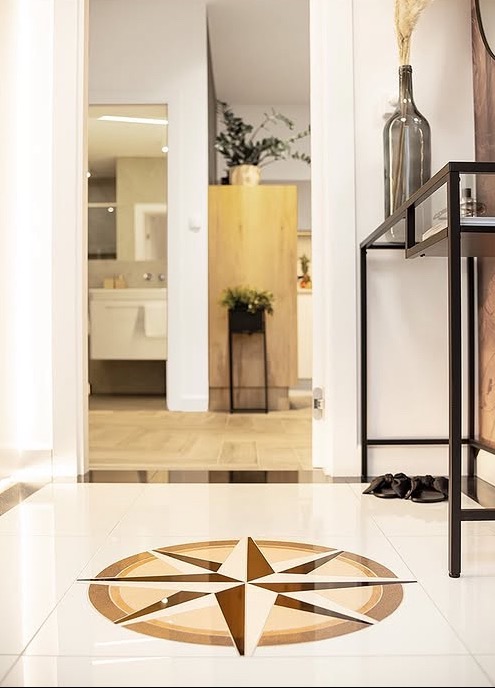 Błyszcząca posadzka z wzorem kompasu w holu. W tle widoczna łazienka i rośliny. Na pierwszym planie czarny stolik konsola z wazonem i suszem.