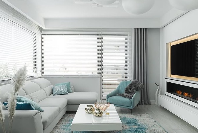 Nowoczesny salon w bieli i szarości z akcentami turkusu: sofa narożna, fotel, stolik kawowy, żaluzje okienne i dekoracyjny kominek elektryczny. Subtelne oświetlenie.