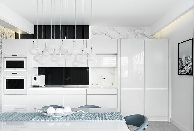 Biała, nowoczesna kuchnia z połyskiem, marmurowym akcentem i designerską lampą. Widoczny stół z dekoracją. Minimalistyczny design z wbudowanym sprzętem AGD.