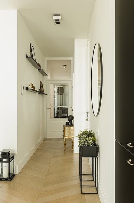 Jasny korytarz z parkietem ułożonym w jodełkę. Na ścianach półki z dekoracjami, lustro w czarnej ramie i złota konsola. Nowoczesne oświetlenie sufitowe.