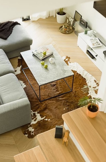 Stylowy salon z szarą sofą, marmurowym stolikiem kawowym i dywanem w krowie łaty. Widok z góry ukazuje nowoczesne wnętrze z elementami dekoracyjnymi i roślinami.