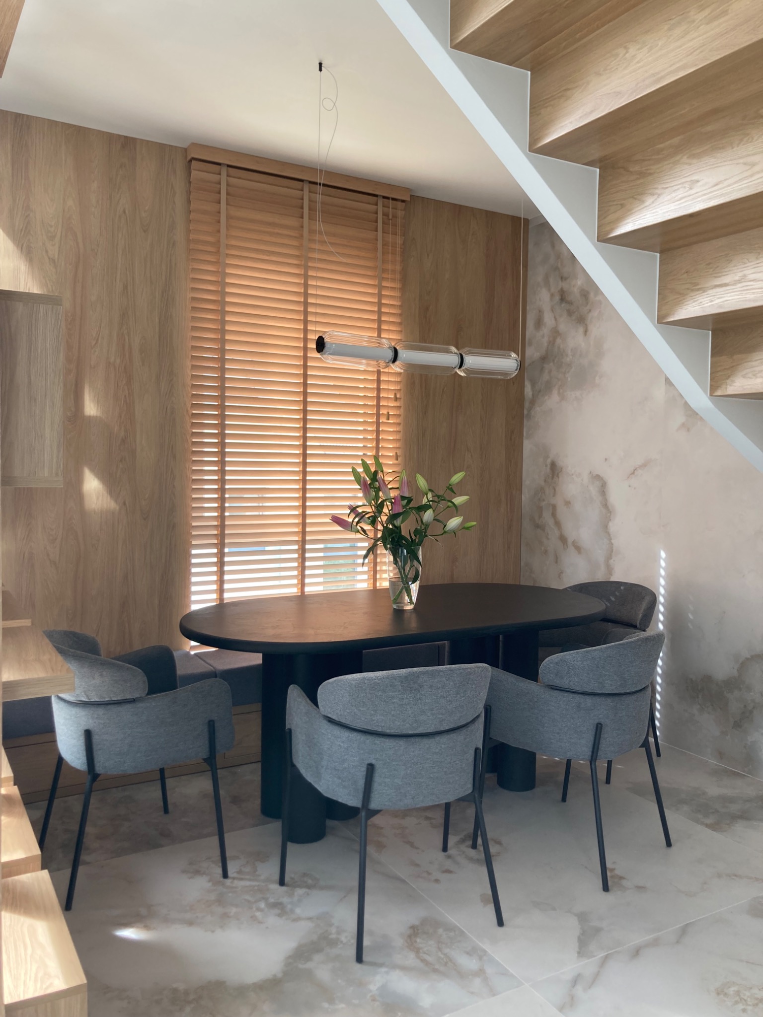 Nowoczesna jadalnia: ciemny, owalny stół otoczony szarymi krzesłami na tle drewnianej ściany z żaluzjami i marmurowej ściany ze schodami.