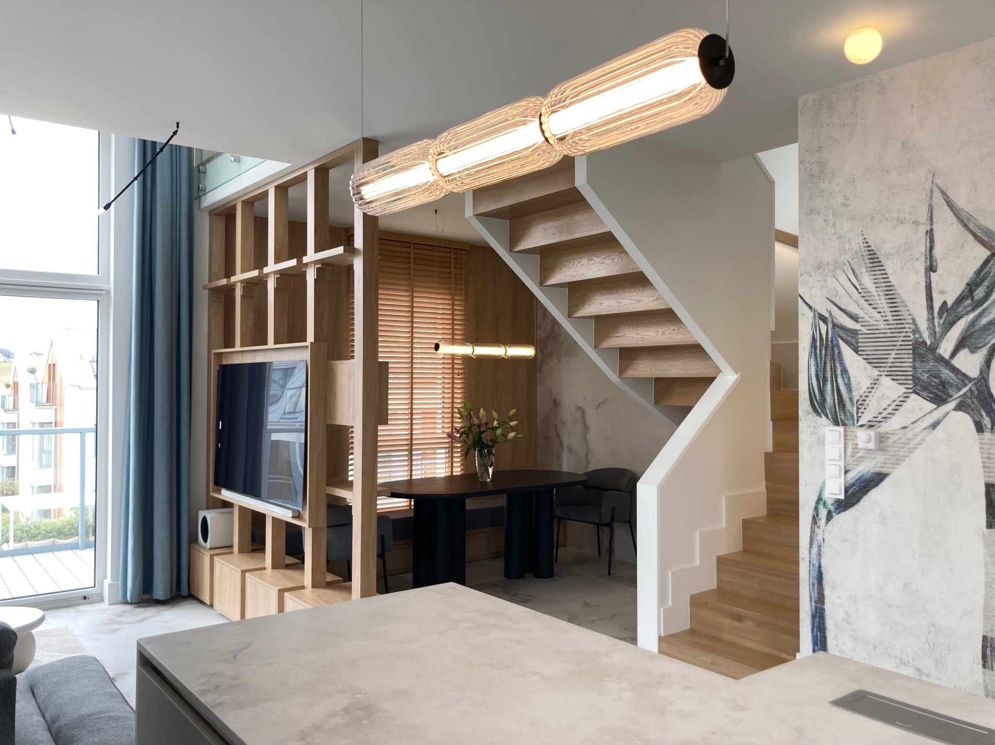 Nowoczesne wnętrze: drewniane schody, stół z kwiatami, ażurowy regał z TV, lampa tubowa. Minimalistyczny design z akcentem na naturalne materiały i jasne kolory.