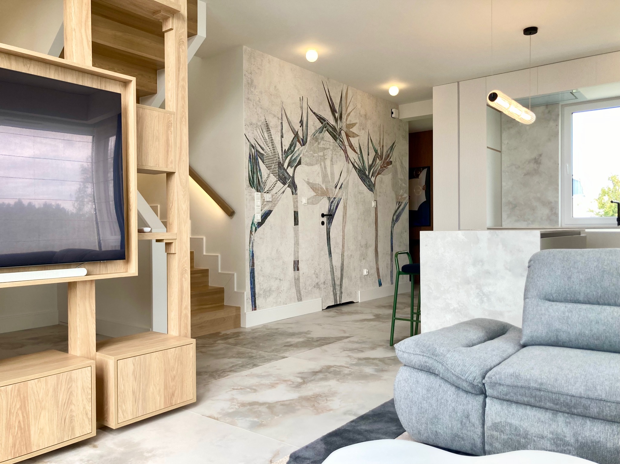 Jasne wnętrze z drewnianą zabudową TV i schodami. Na ścianie dekoracyjny motyw roślinny, szara sofa i marmurowe płytki podłogowe.