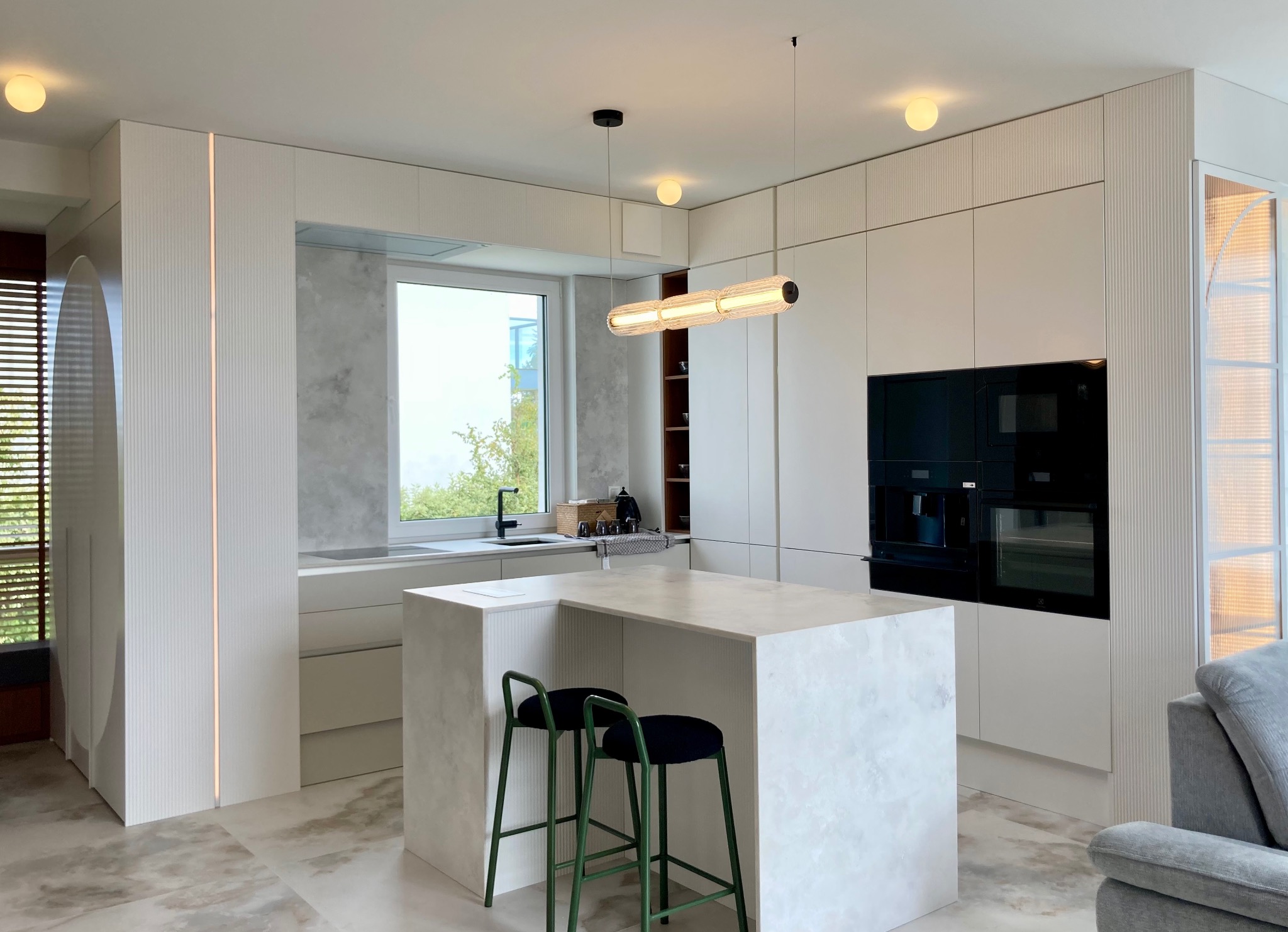 Nowoczesna kuchnia w jasnych barwach z wyspą i zielonymi hokerami, oświetlona podłużną lampą. Minimalistyczny design, zabudowa kuchenna z teksturą.