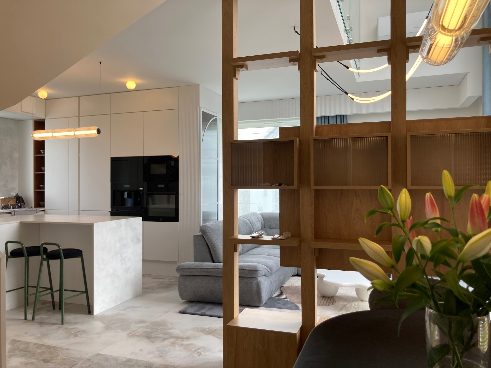 Nowoczesne wnętrze: biała kuchnia z wyspą, szara sofa i drewniany ażurowy regał. Minimalistyczny design, jasne kolory, kwiaty w wazonie na pierwszym planie.