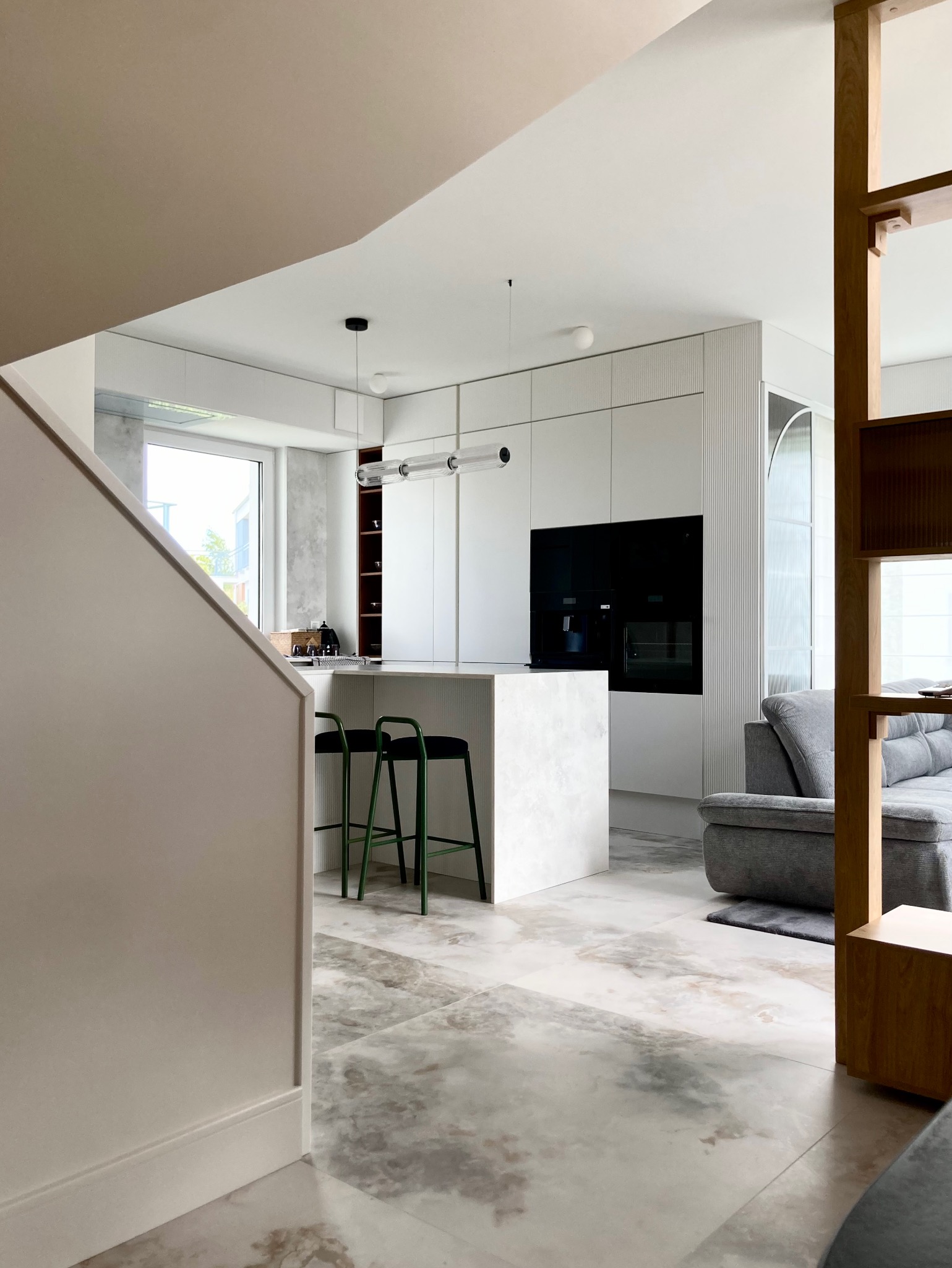 Nowoczesna, otwarta kuchnia z wyspą i zielonymi hokerami. Marmurowe płytki podłogowe, białe szafki z frezowanymi frontami i czarny sprzęt AGD w zabudowie.