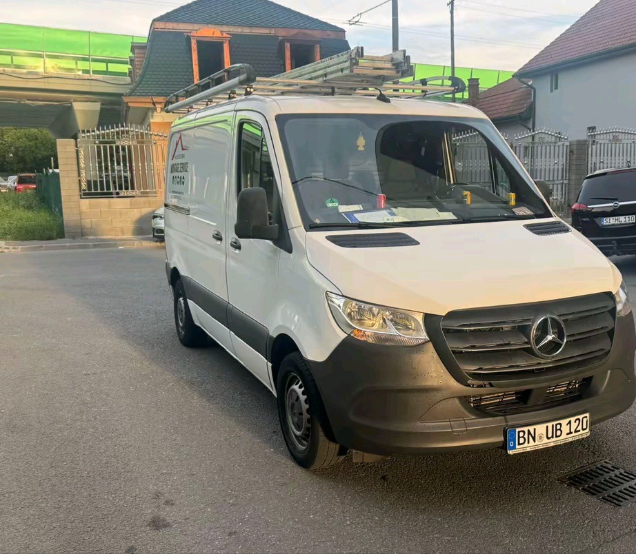 Biały van Mercedes z drabina na dachu, zaparkowany na ulicy, z widocznym logo firmy dekarskiej na boku i zielonym dachem budynku w tle. Tablica rejestracyjna widoczna.