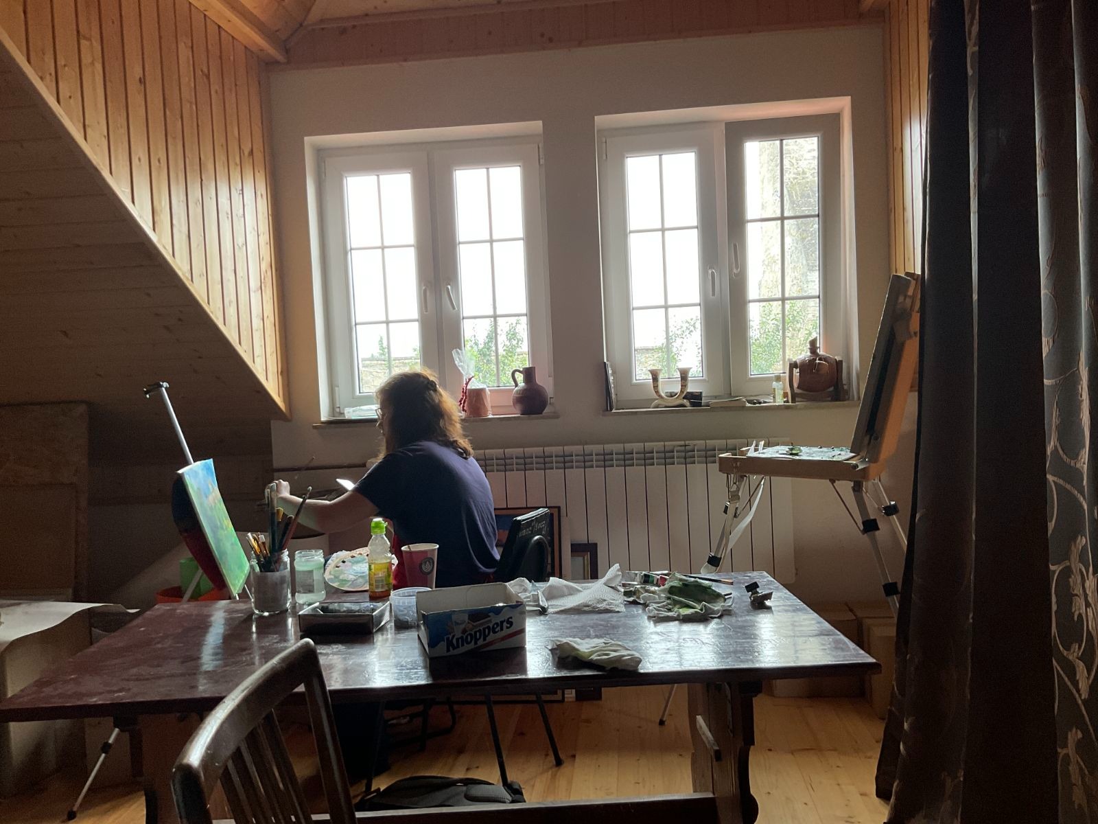 Artystka maluje obraz na sztaludze w pracowni na poddaszu, z oknami i narzędziami malarskimi na drewnianym stole. Widoczne farby, pędzle i szkic.