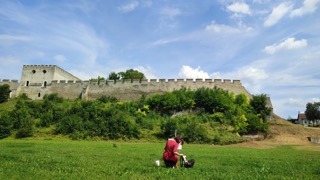 Artysta w stroju historycznym maluje zamek na wzgórzu w plenerze. Widok z dołu na zielone pole i błękitne niebo z chmurami.