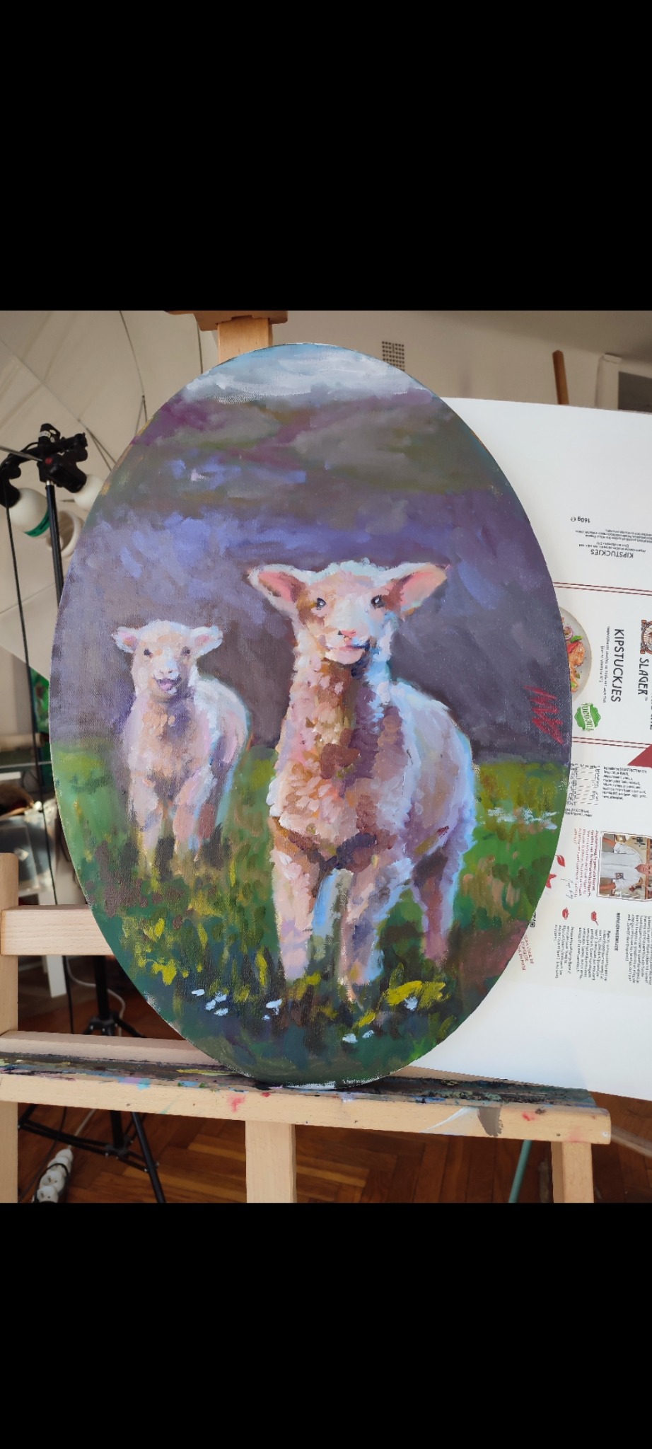 Owalny obraz olejny przedstawia dwa jagnięta na łące, namalowane ekspresyjnymi pociągnięciami pędzla. Widoczne na sztaludze w pracowni artystycznej.