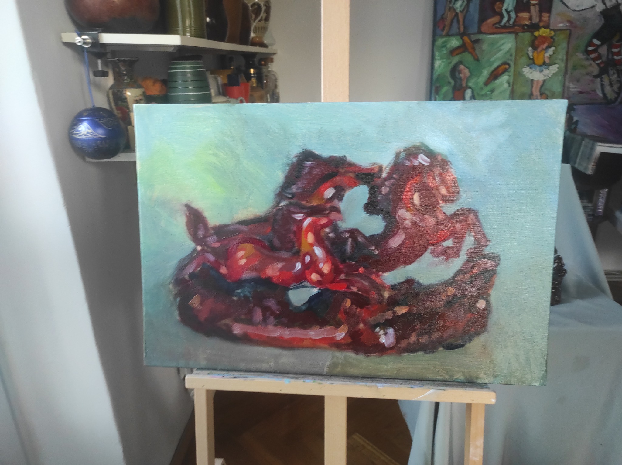 Obraz olejny na płótnie przedstawia dynamiczną scenę z dwoma brązowo-czerwonymi końmi na tle błękitno-zielonym. Obraz stoi na drewnianym sztaludze, w tle półka z bibelotami.