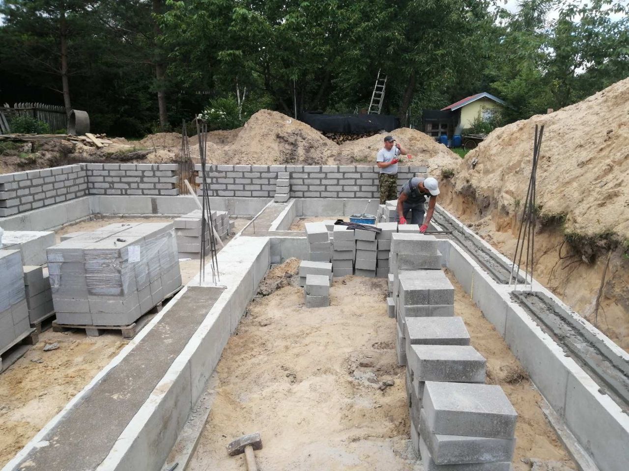 Budowa fundamentów: pracownik układa bloczki betonowe na fundamencie. Widoczne zbrojenia, paleta z bloczkami i narzędzia na placu budowy.