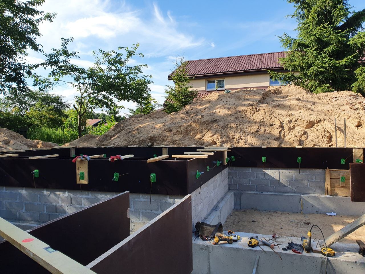 Fundamenty domu w trakcie budowy: szalunki z desek, bloczki betonowe, narzędzia budowlane rozłożone na fundamencie, w tle hałda piasku i dom z czerwonym dachem.