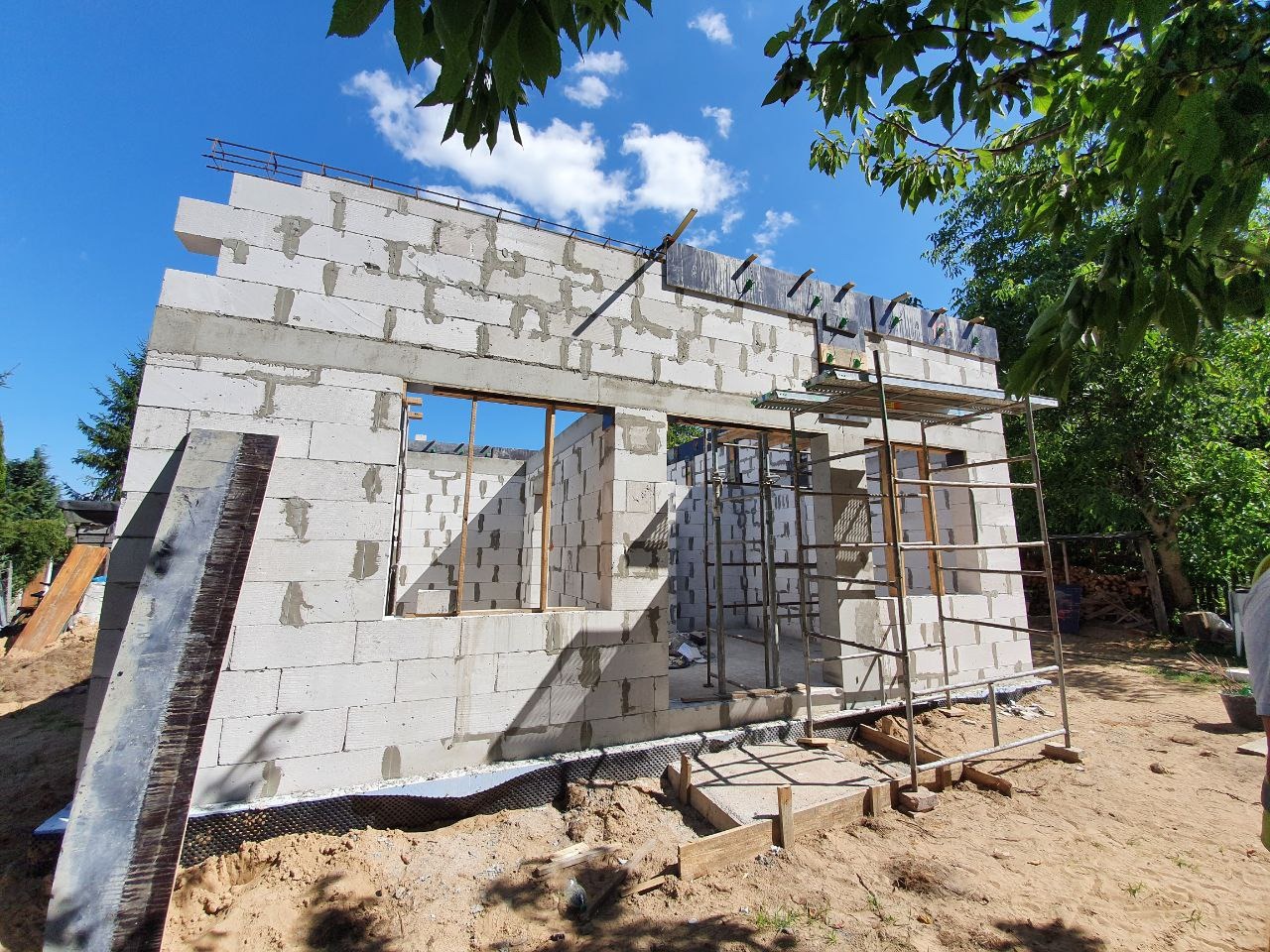 Wznoszenie ścian z betonu komórkowego na fundamencie. Widoczny szalunek okienny, rusztowanie i zbrojenie. Prace budowlane w słoneczny dzień.