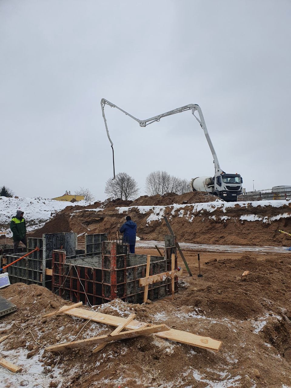 Zimowa budowa fundamentów: szalunek zbrojony, pracownicy i betonowóz z pompą na placu budowy. Śnieg i ziemia w tle.