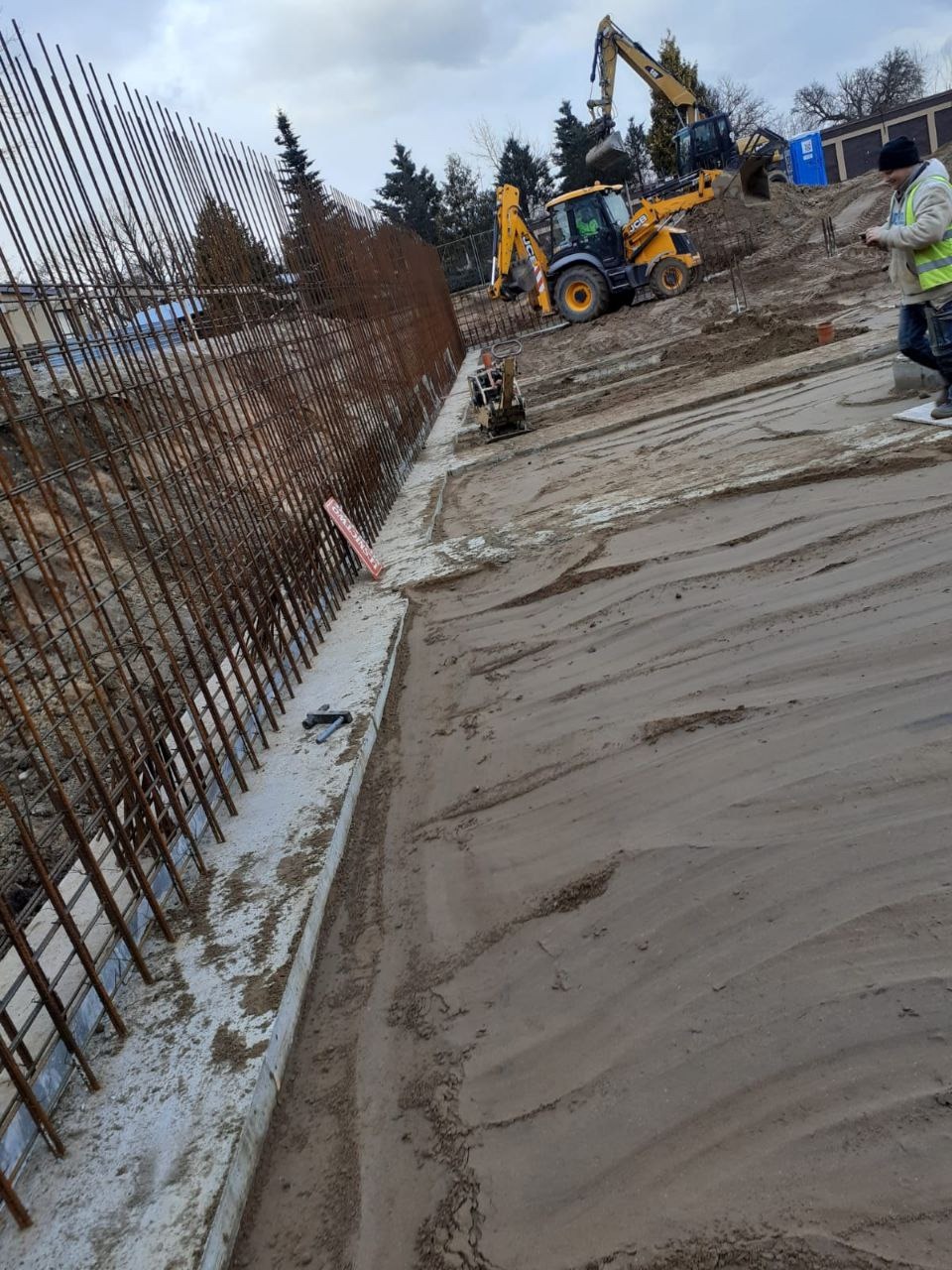 Zbrojenie fundamentów pod budowę, widoczne pręty stalowe i betonowy wieniec. W tle koparki i pracownik budowlany. Teren przygotowany pod wylewkę.