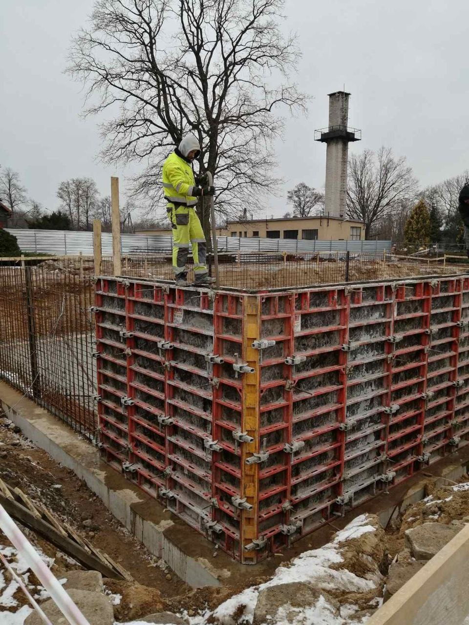 Pracownik w odzieży roboczej stoi na szalunku fundamentu budynku w trakcie budowy, widoczny zbrojony fundament i drzewo bez liści w tle.